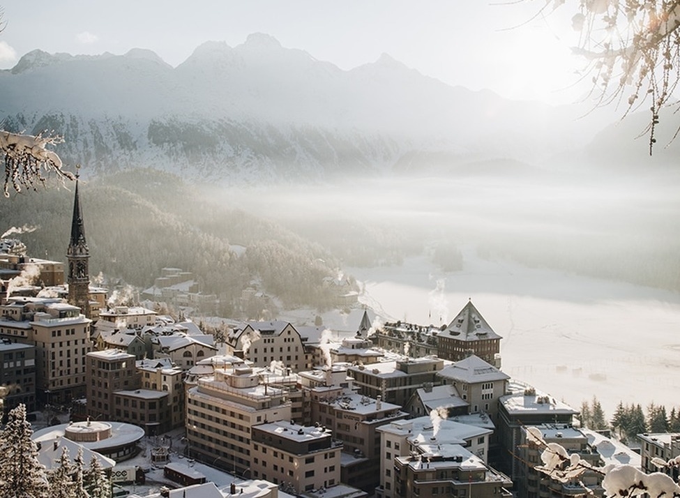 Snowy rooftops of Badrutt’s Palace in St. Moritz overlooking a mist-covered alpine valley and frozen lake.