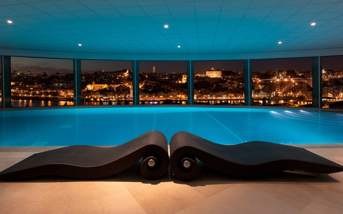 Loungers beside an indoor pool, with the after dark city in the background.