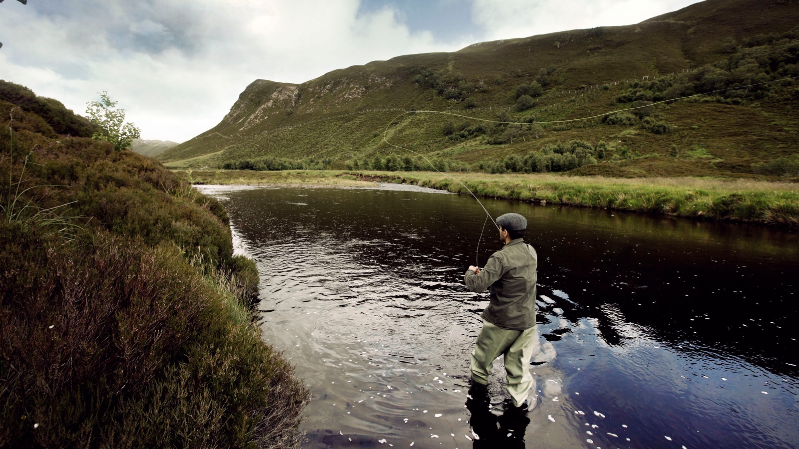 A man fly fishing in a river surrounded by rolling green hills under an overcast sky.