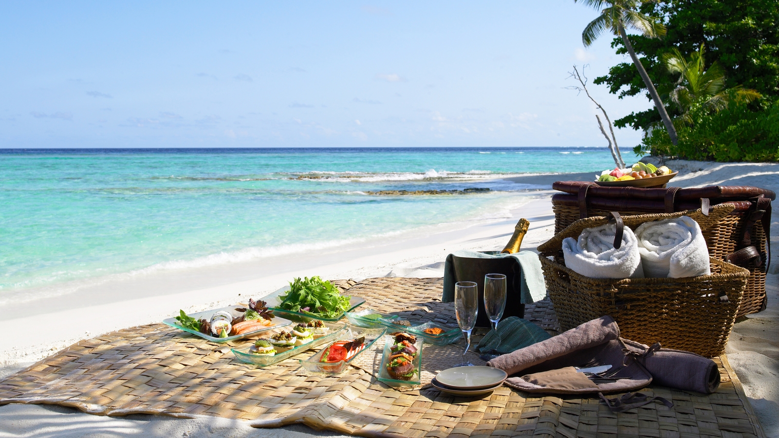 Gourmet picnic spread on a straw mat at a white sand beach with turquoise water and palm trees.