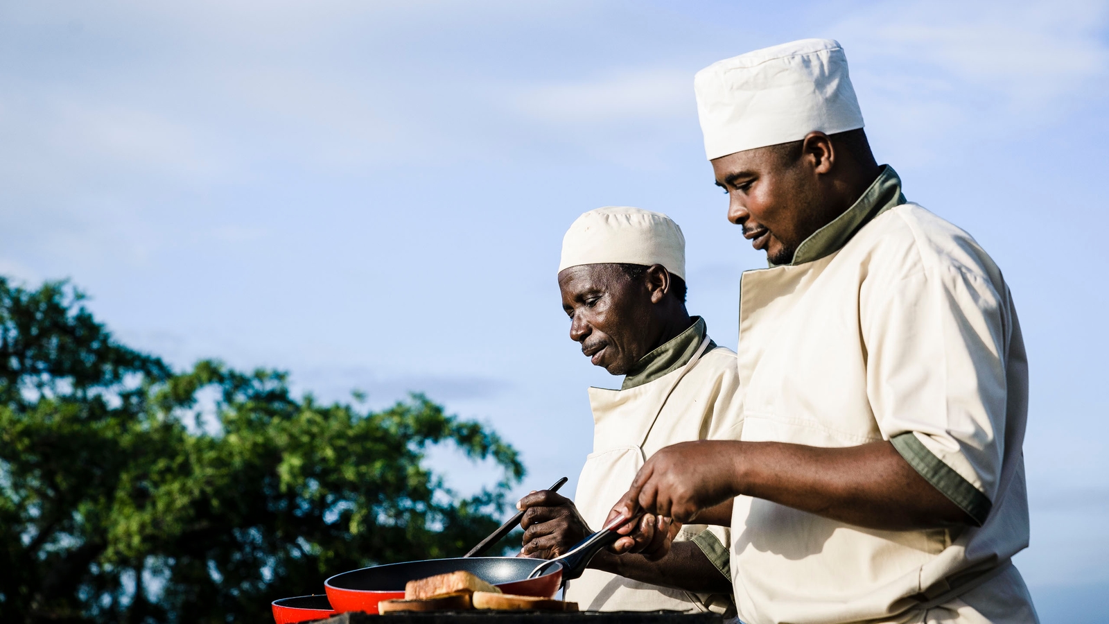 Two men in chef's hats and jackets cooking in red frying pans outside