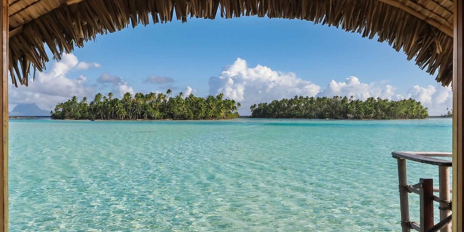 View of a blue lagoon and green islet framed by a thatched-roof overwater bungalow at Le Taha'a by Pearl Resorts in French Polynesia.