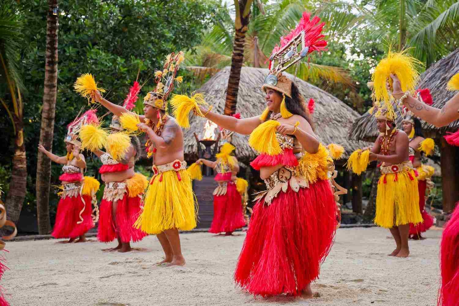 Dancers in traditional red and yellow Polynesian attire performing on a sandy beach at Le Taha'a by Pearl Resorts in French Polynesia.