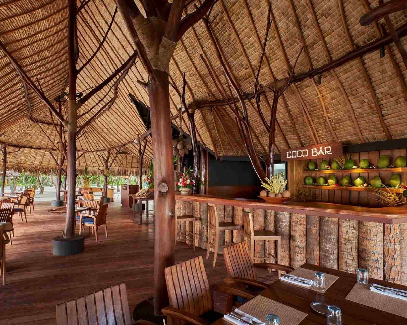 Open-air restaurant pavilion with a thatched wooden roof, rustic bar, and dining tables at Le Taha'a by Pearl Resorts in French Polynesia.