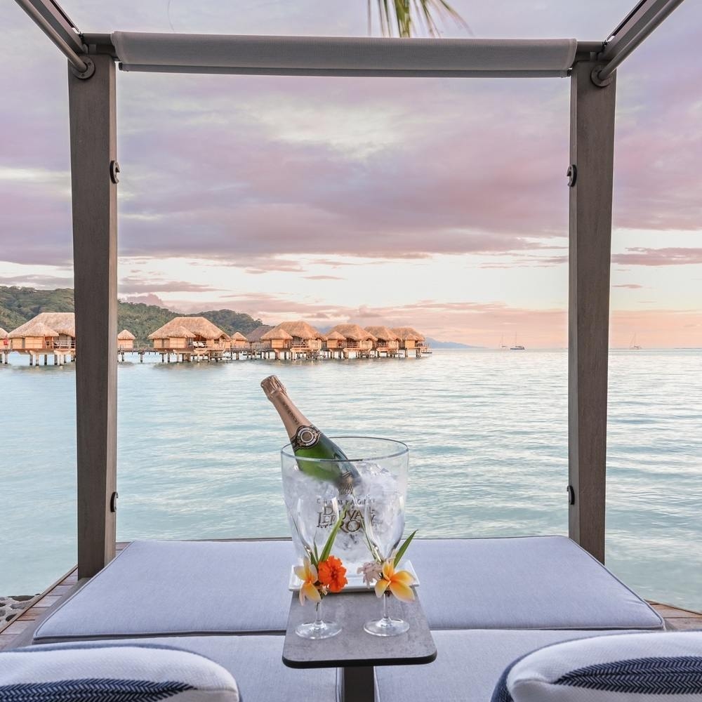 Champagne bucket on a cushioned daybed framing overwater bungalows and a tranquil lagoon at Le Taha'a by Pearl Resorts in French Polynesia.