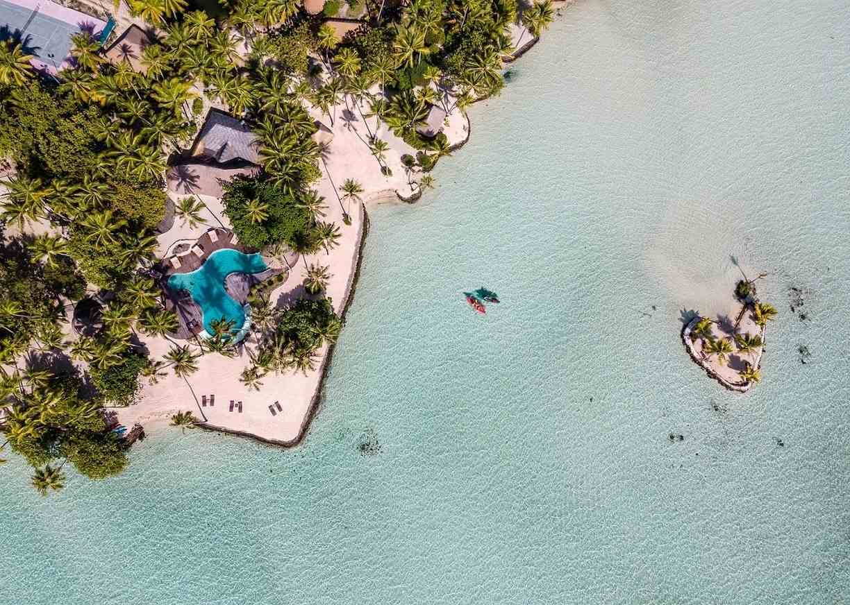 Overhead view of a swimming pool and sandy beach surrounded by palm trees and shallow turquoise water at Le Taha'a by Pearl Resorts in French Polynesia.