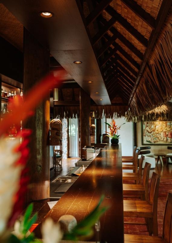 Polished wooden bar counter and stools inside a resort lounge with a thatched roof at Le Taha'a by Pearl Resorts in French Polynesia.