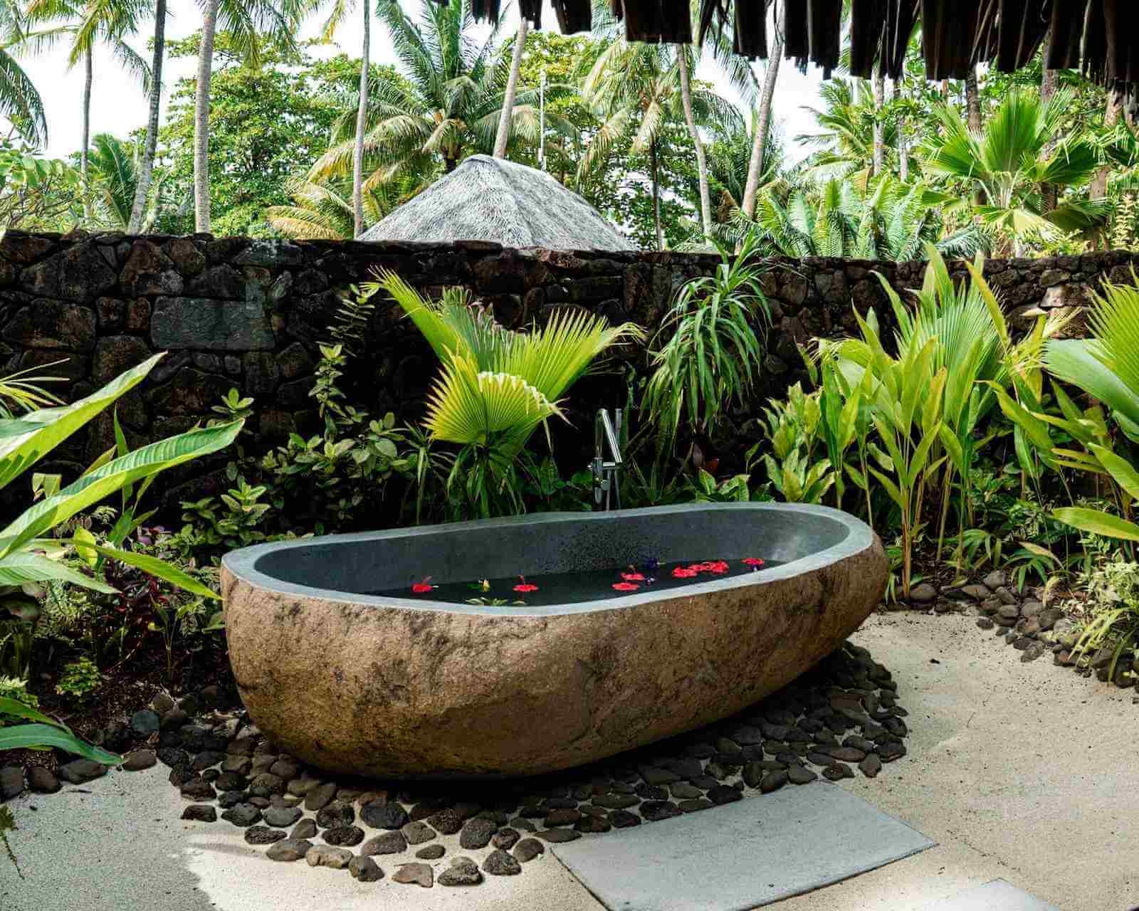 Carved stone bathtub filled with water and floating flowers outdoors at Le Taha'a by Pearl Resorts in French Polynesia.