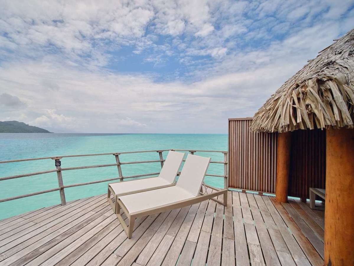 Two sunbeds on a wooden overwater terrace overlooking a bright turquoise lagoon at Le Taha'a by Pearl Resorts in French Polynesia.