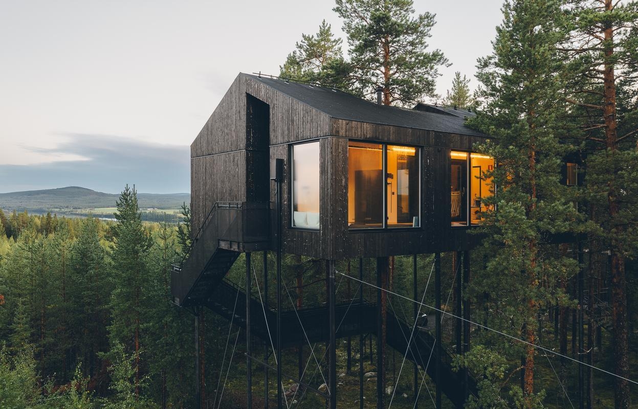 A large black wooden structure on high stilts surrounded by trees under a grey sky.