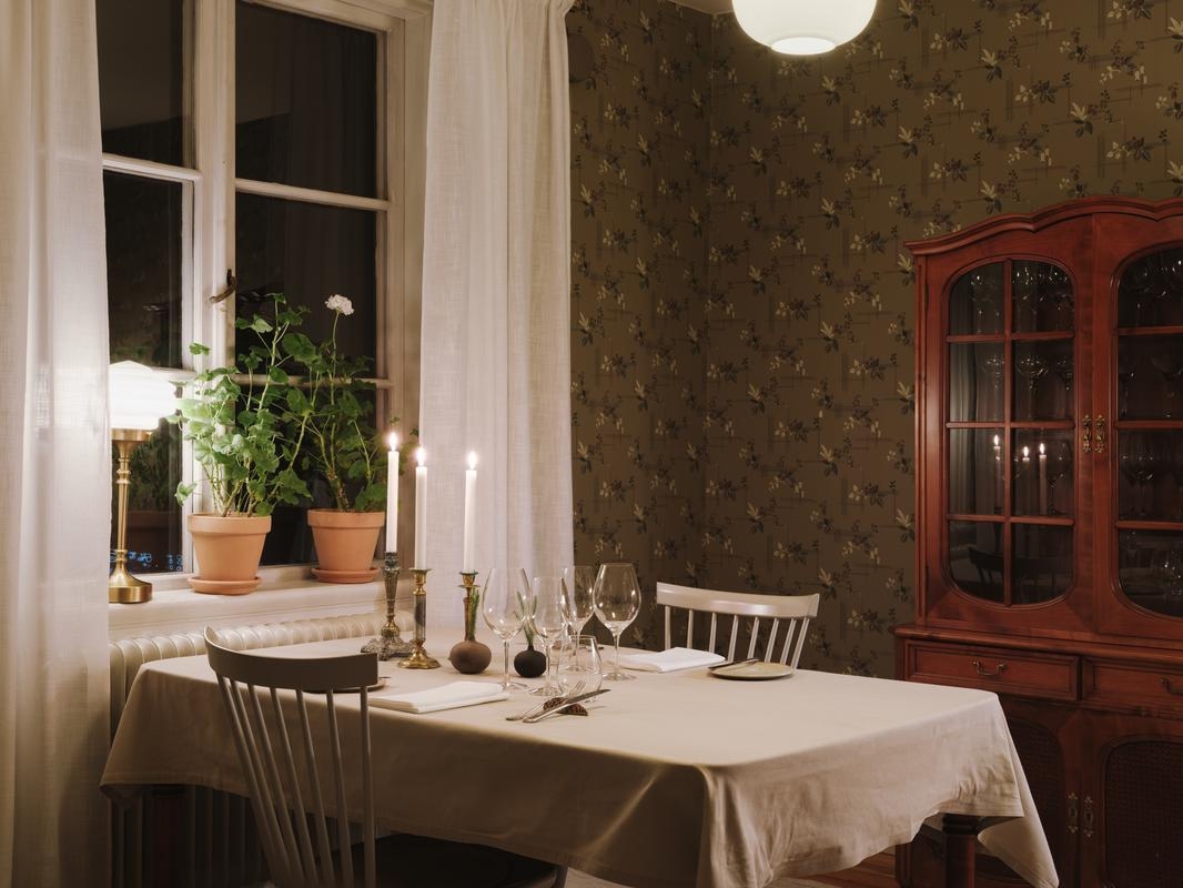 A set dinner table with candles and wine glasses in a room with floral wallpaper and a wooden cabinet.