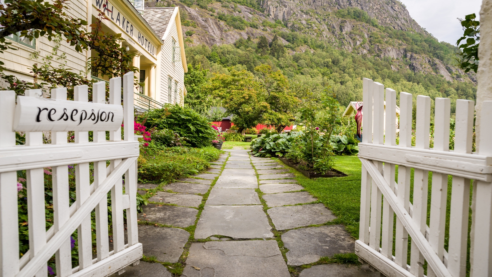 A pathway leading to a lush garden with "resepsion" sign on a white gate, near a hotel, with mountains in the background.