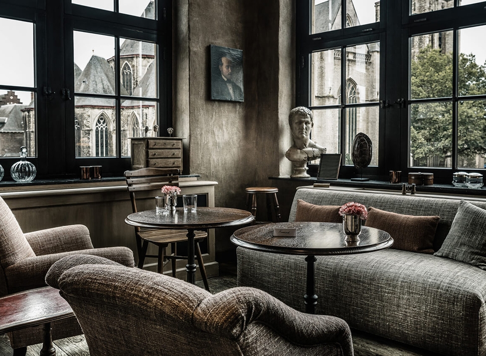 A dark, moody lounge with vintage armchairs, wooden tables, a bust statue, and tall windows looking out at a gothic church.