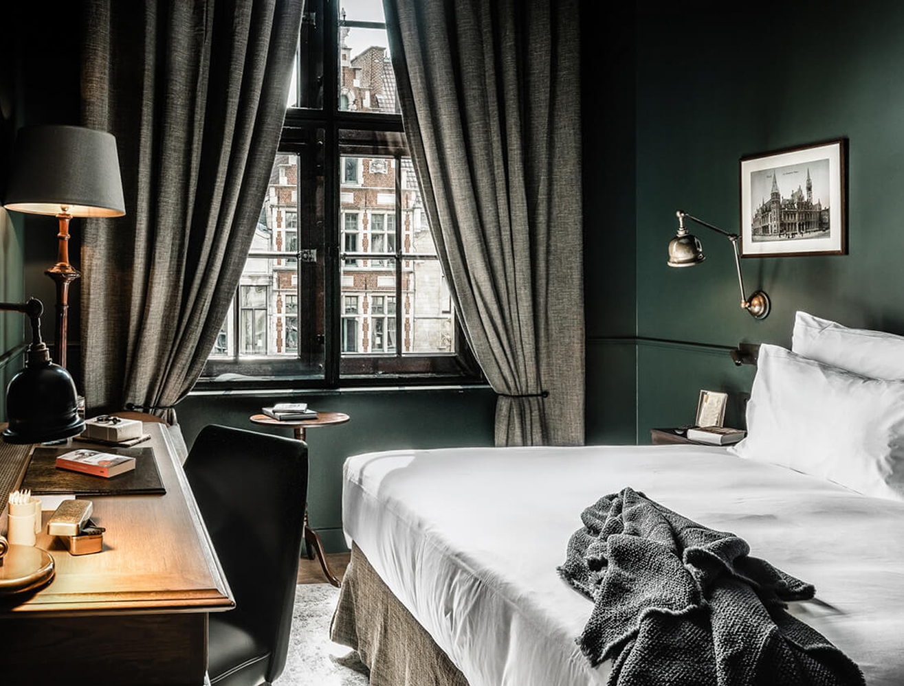 A bedroom with dark green walls, a white bed, a desk, and a window with curtains looking out at historic brick buildings.