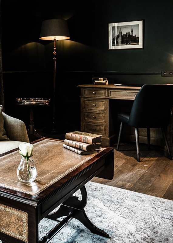 A moody lounge with dark walls, a wooden desk, and a leather-topped coffee table holding antique books and a white rose.