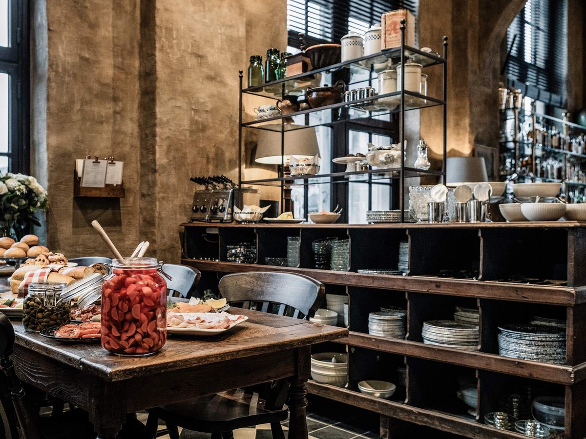 A rustic breakfast buffet on a wooden table with bread and a large jar of pickled vegetables, alongside shelves of plates.