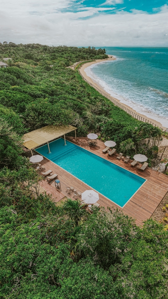 A top-down view of a pool next to a beach, flanked by lush greenery.