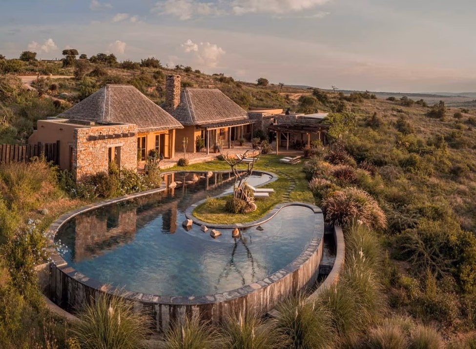A stone luxury villa with a large, uniquely shaped infinity pool overlooking a wide, sunlit savanna landscape.