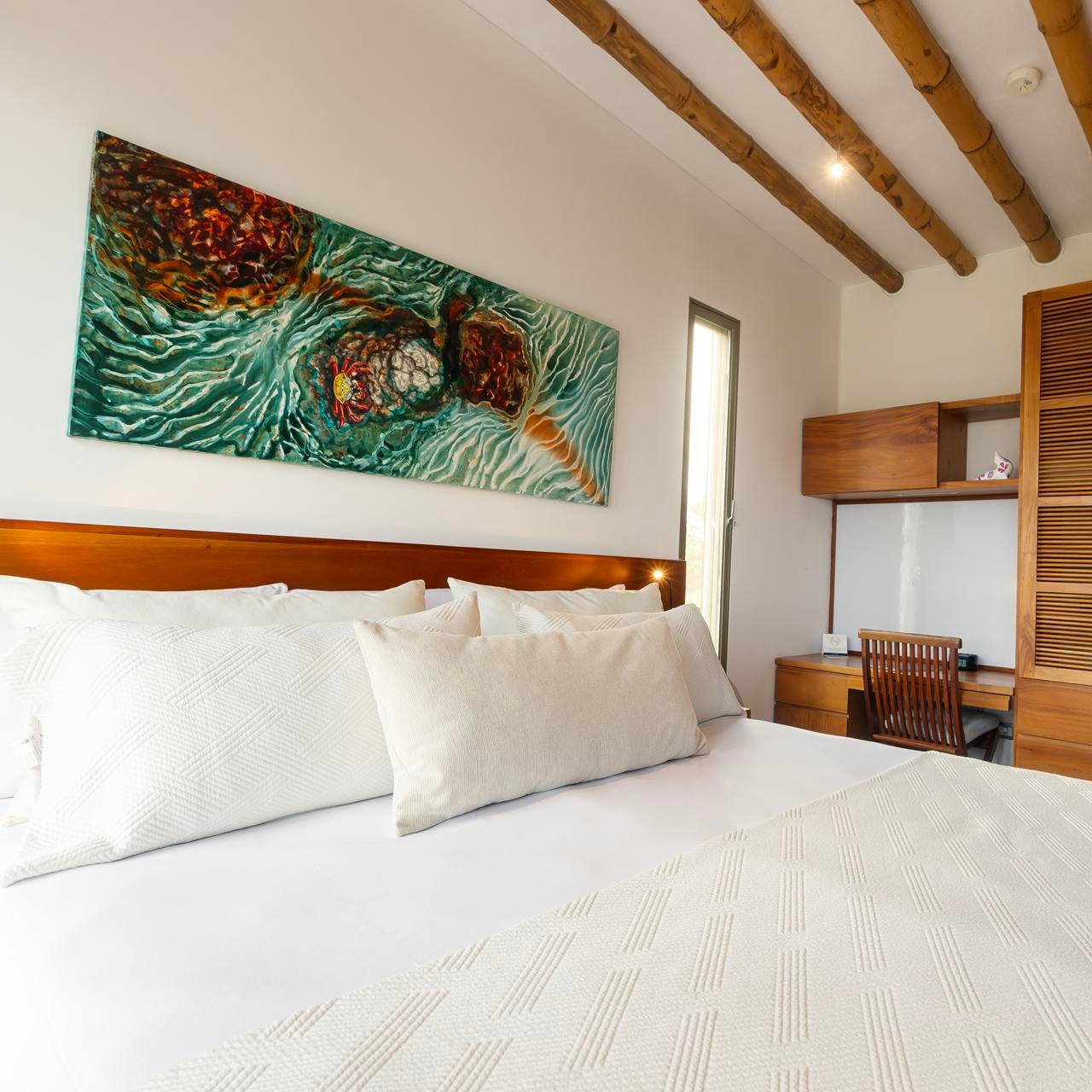 Modern bedroom with a long abstract painting above the bed, exposed wooden beams, and soft lighting.