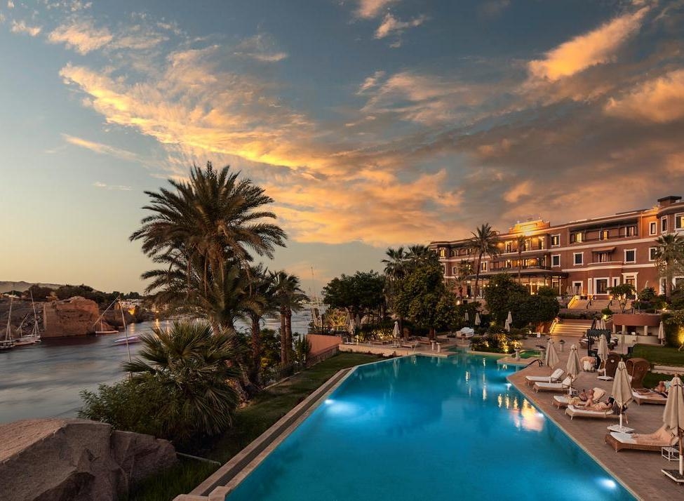 A luxury hotel with a bright blue infinity pool and palm trees under a dramatic sunset sky in Aswan.