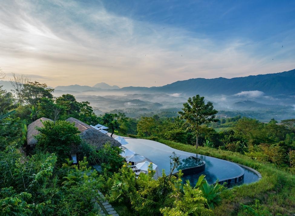 The infinity pool at Avana Retreat overlooking a misty valley and mountain range with thatched-roof villas and tropical greenery.