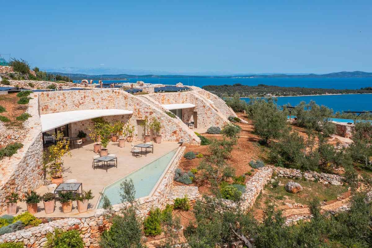 High-angle view of a modern stone villa built into a green hillside overlooking the ocean.