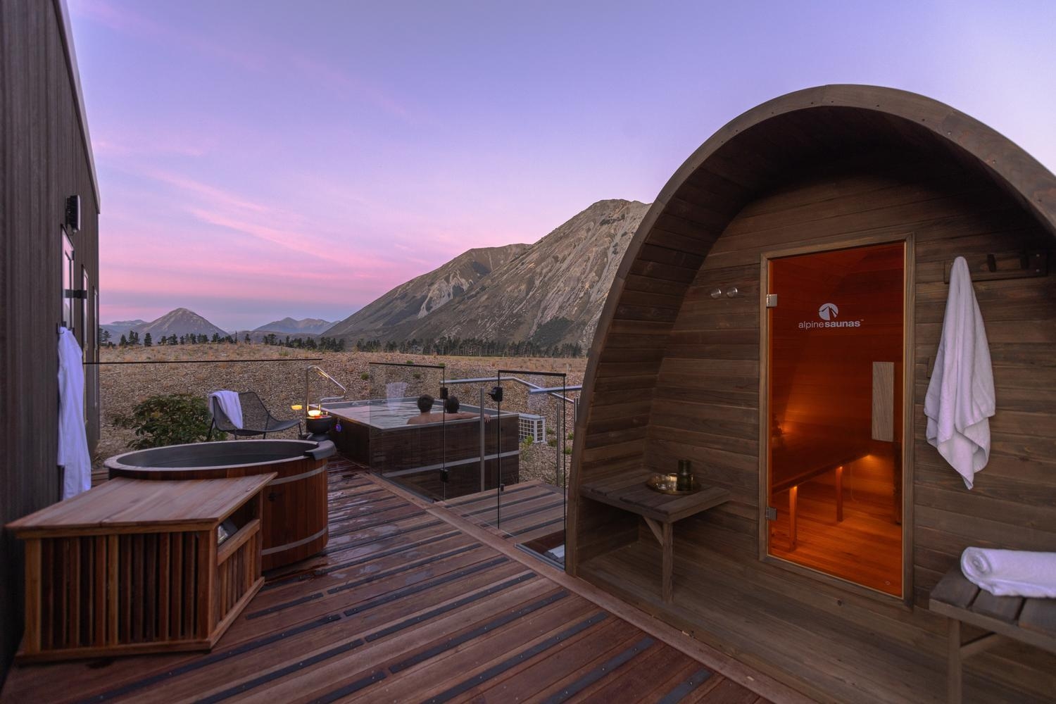 Wooden spa deck with a hot tub, cold plunge, and an alpine saunas barrel sauna overlooking mountains during sunset.