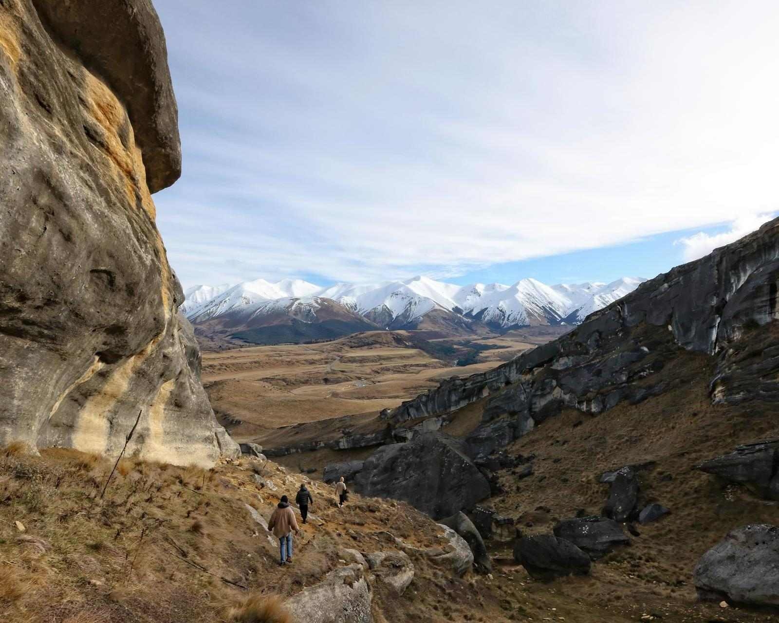 People hiking down a rocky trail surrounded by large rock formations with snow-capped mountains in the distance.