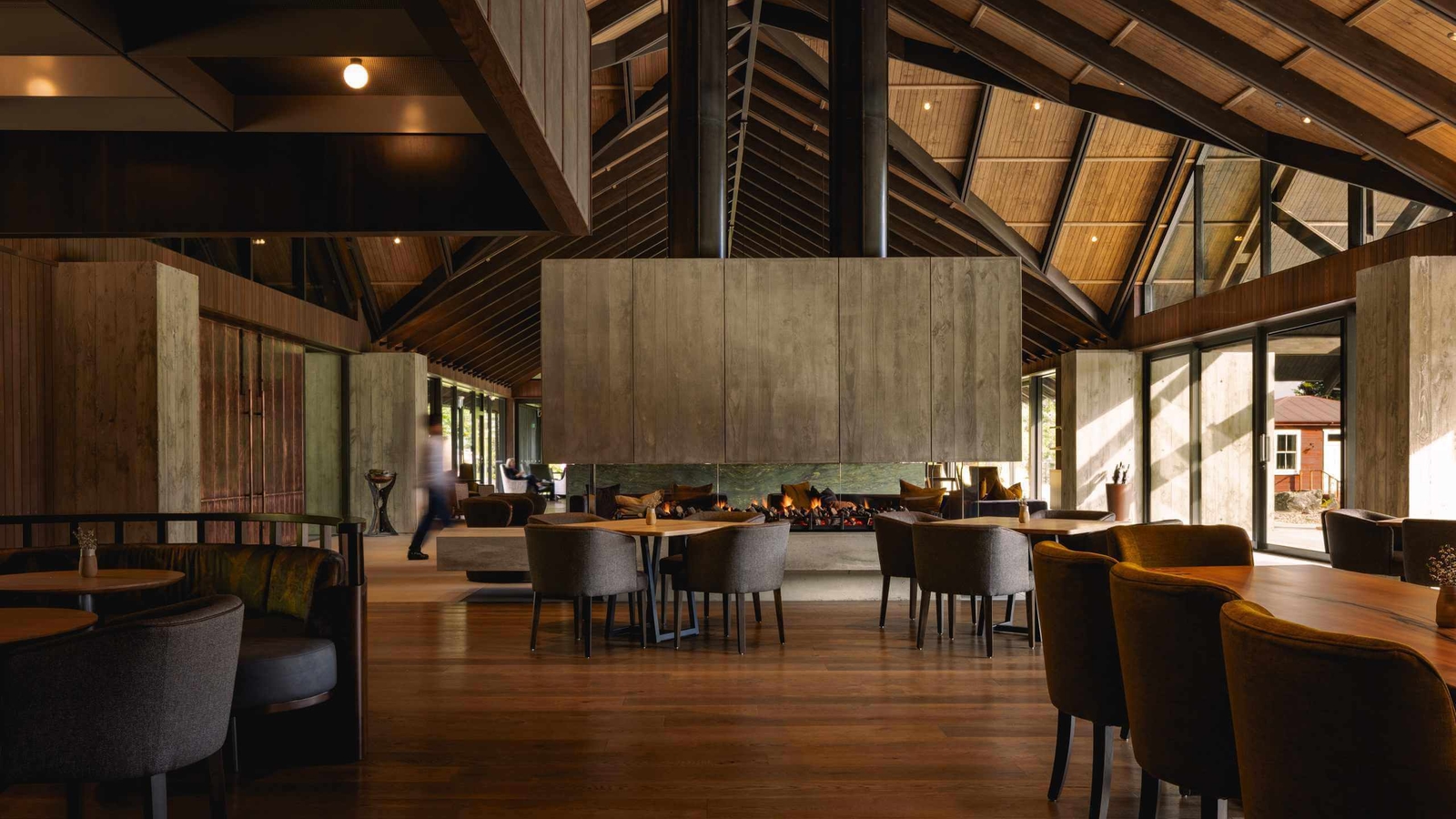 Modern lodge dining hall with wooden vaulted ceilings, dining tables, and a central fireplace.
