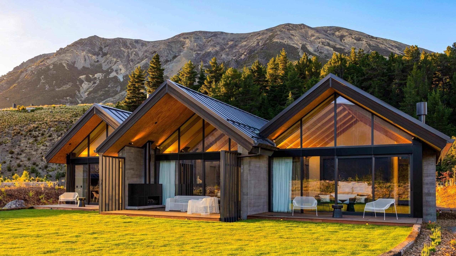 Exterior view of modern gabled villas with glass windows and a lawn before a mountain backdrop.