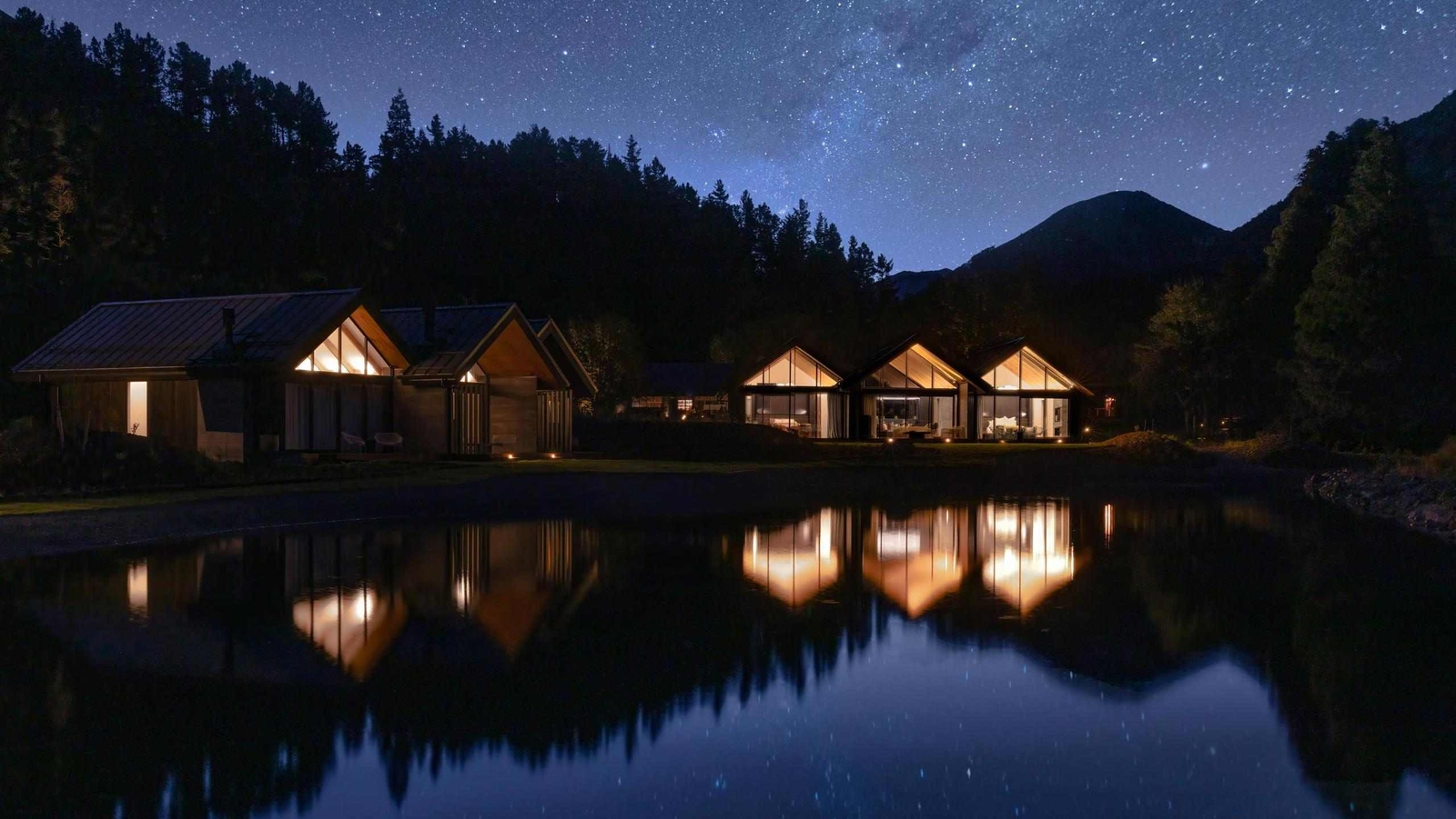 Night view of lit modern villas reflecting in a lake under a starry sky.