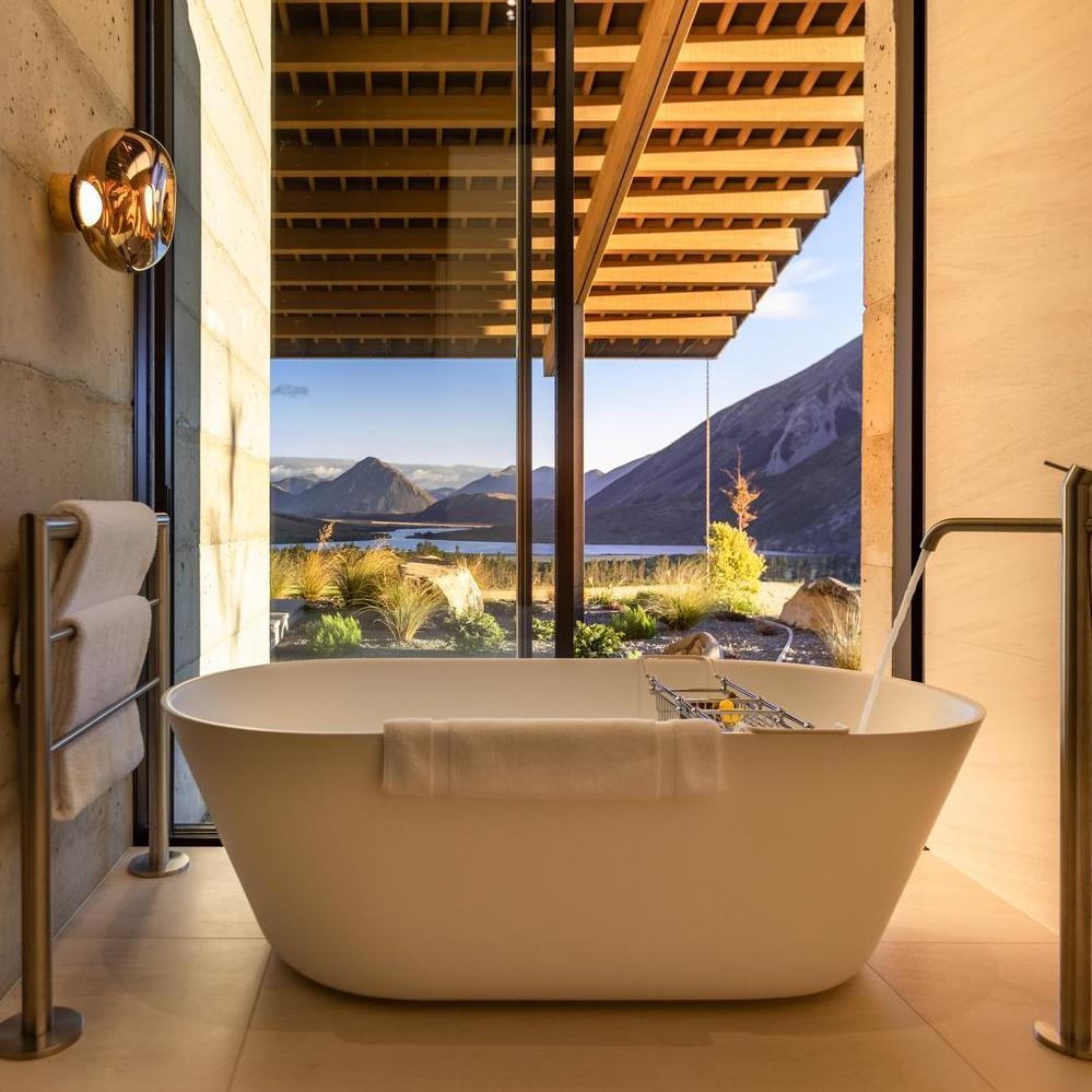 Modern freestanding bathtub inside a luxury bathroom with glass windows overlooking mountains and a lake.