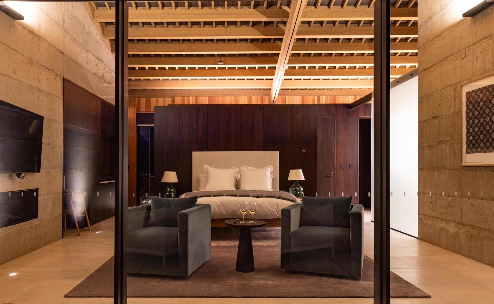 Modern luxury bedroom with wooden exposed ceiling beams, upholstered bed, and armchairs framing a small table.