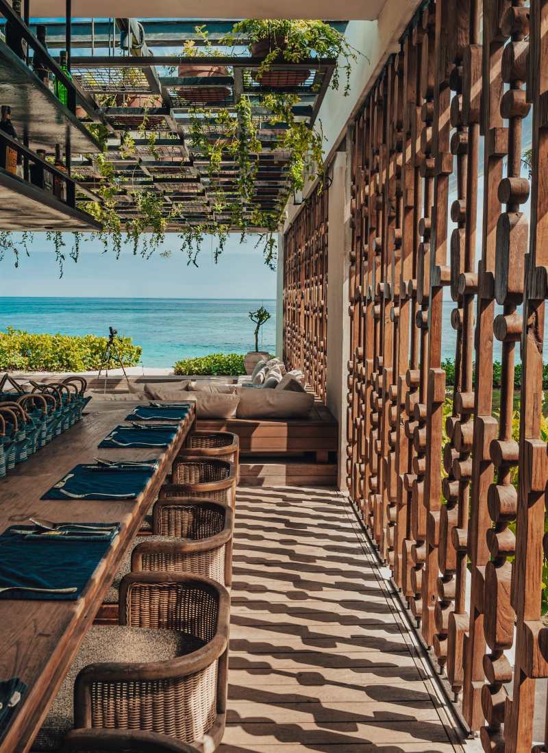 One of Cap Karoso's villa's open-air dining area with a long wooden table, wicker chairs, and a decorative wood screen facing the ocean.