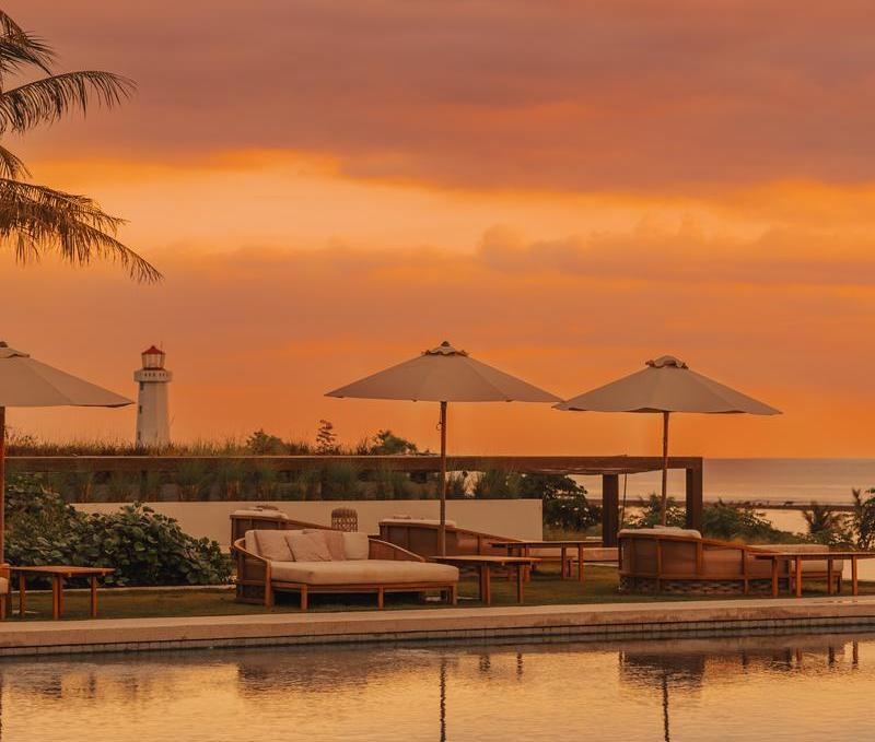 Silhouette of a lighthouse and poolside daybeds at sunset with a warm orange sky reflecting in the water.