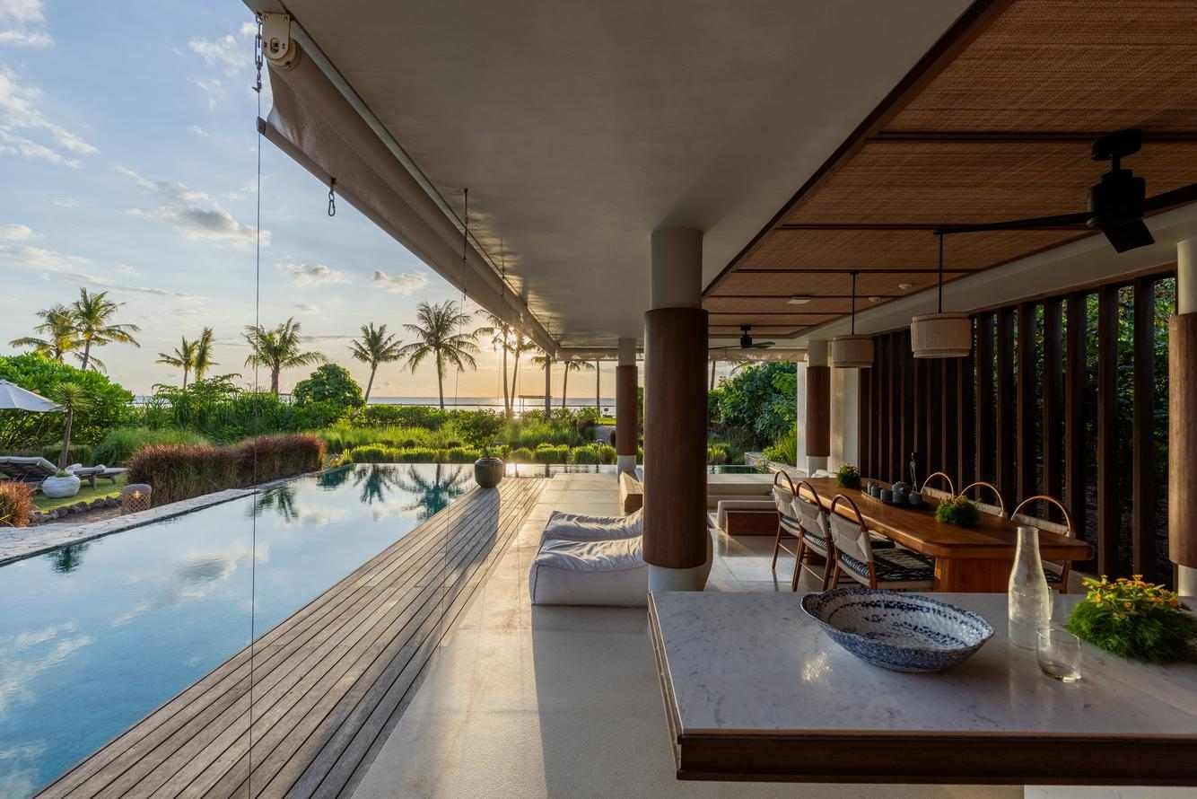 Modern villa at Cap Karoso featuring a terrace with an infinity pool, wooden dining table, and palm trees against a sunset sky.
