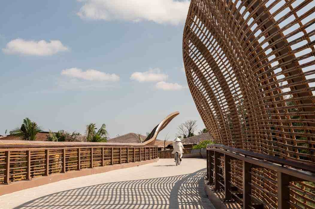 A person wearing a conical hat cycles across a bridge with a large, curved bamboo architectural feature.