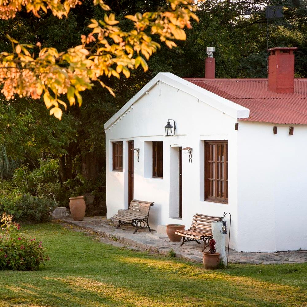 A quaint white cottage with a red tile roof, benches outside, surrounded by greenery.