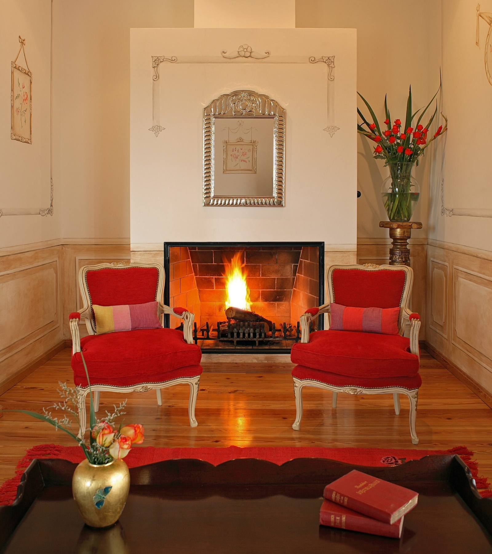 Elegant living room with lit fireplace flanked by red armchairs and decorative mirror above.