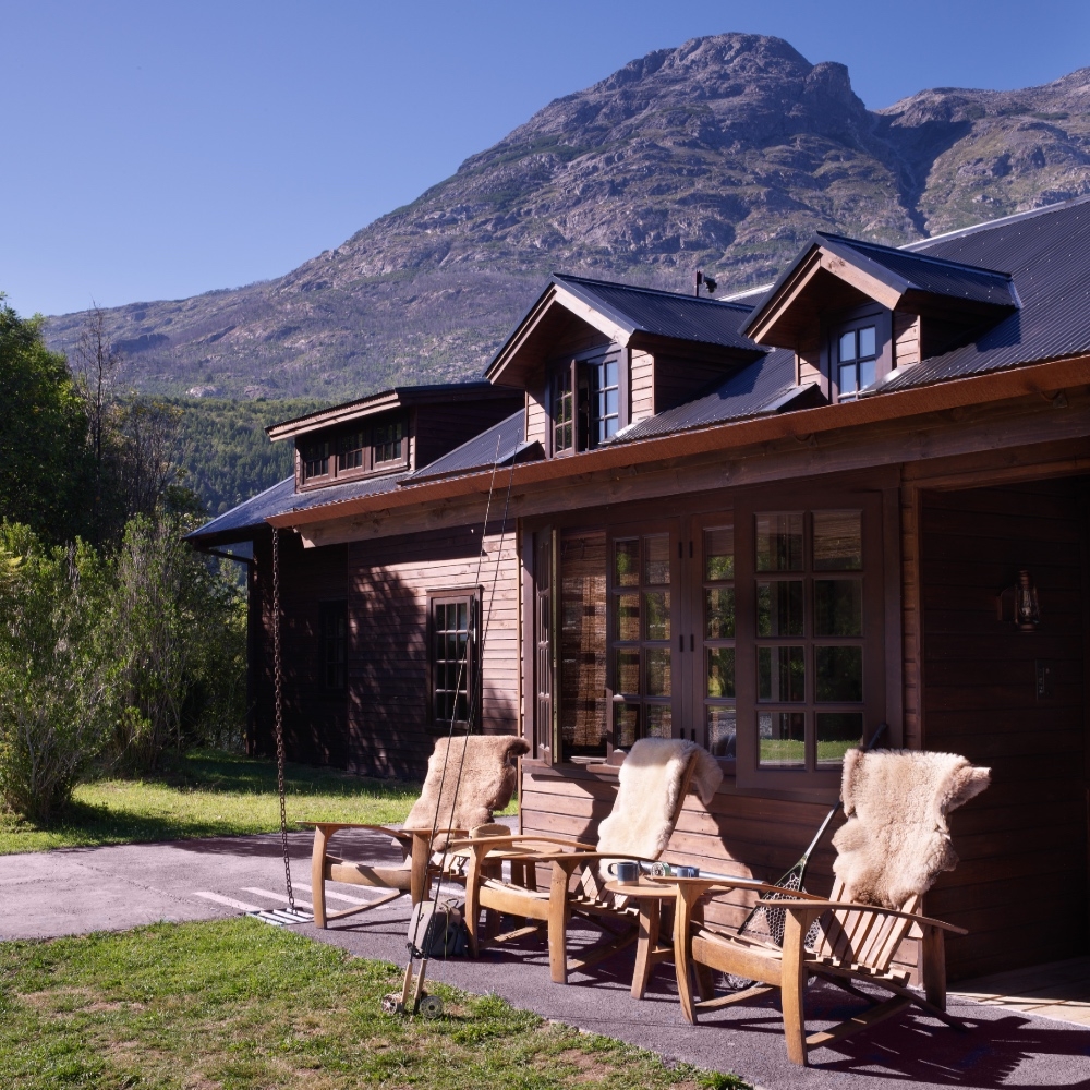 Wooden cabin with chairs in front and mountain backdrop.
