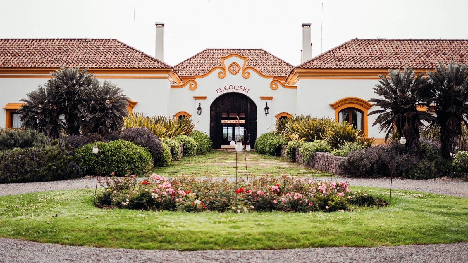 Elegant hacienda-style building "El Colibri" with colorful garden and pathway.