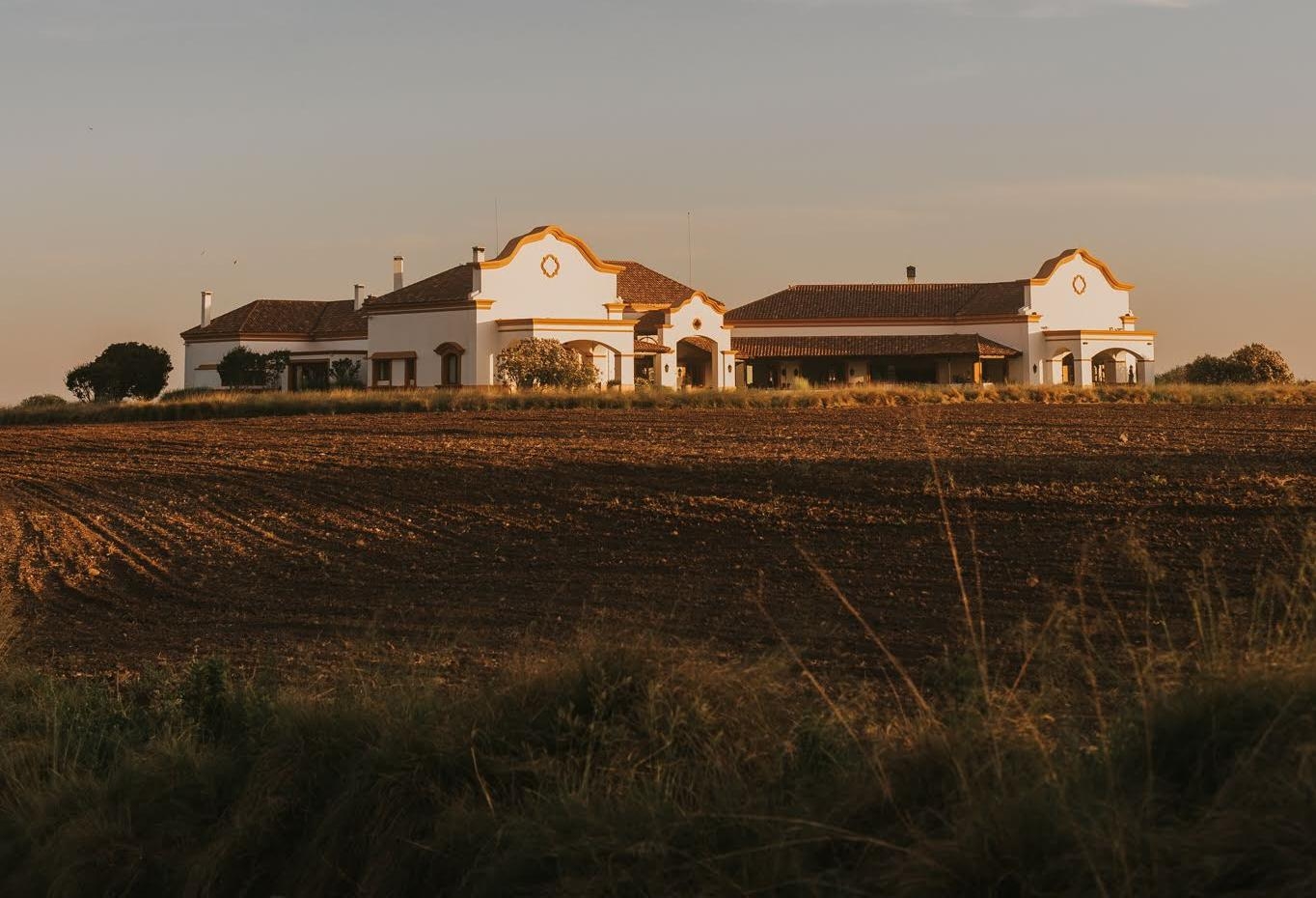 Traditional white farmhouse with terracotta roofs at sunset, surrounded by plowed fields.