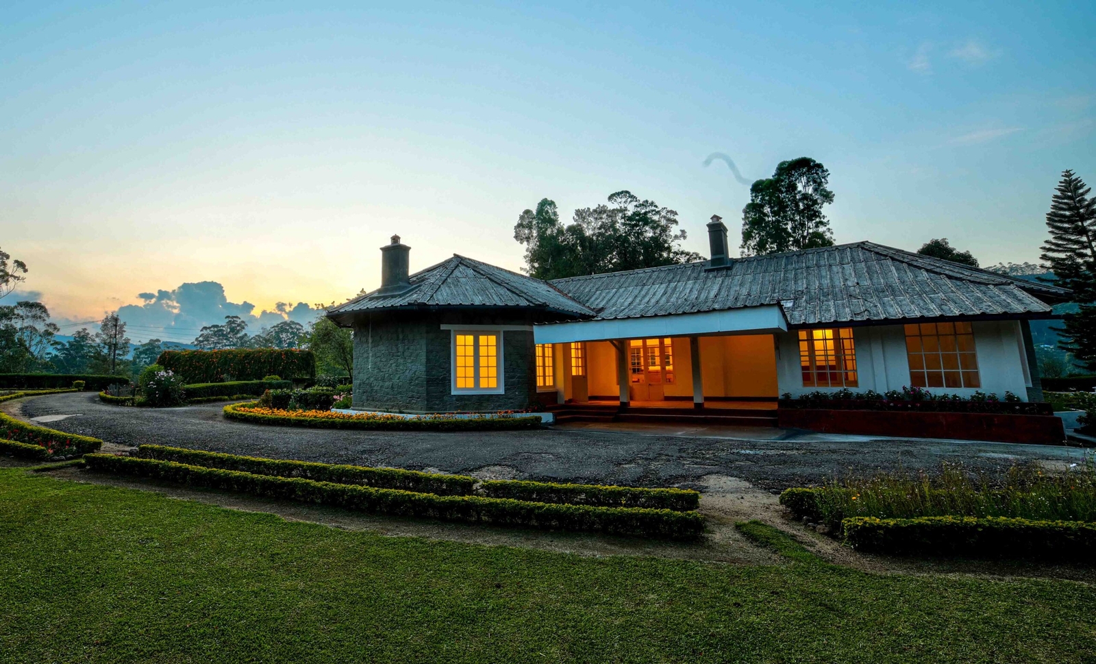 A quaint cottage with lit windows at dusk, surrounded by manicured gardens.