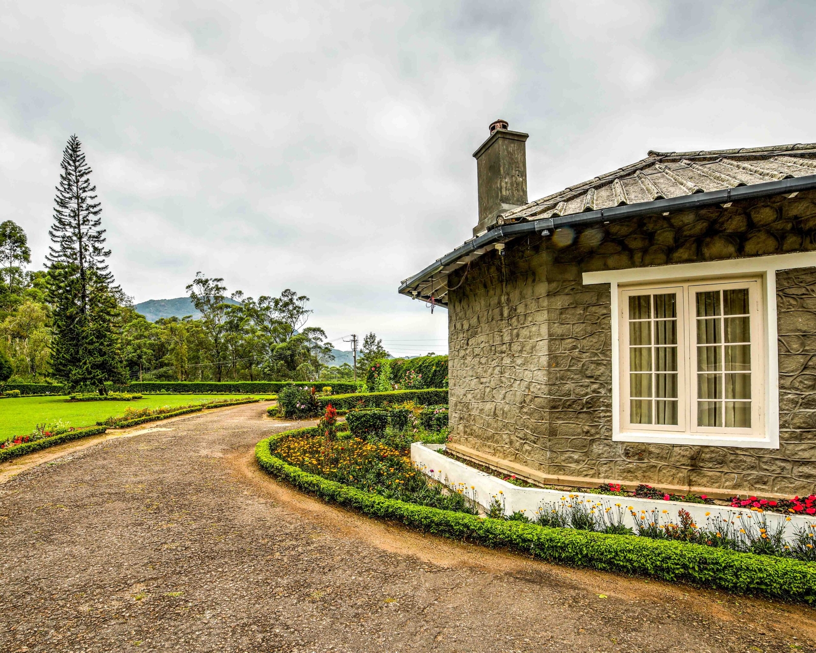 Stone cottage with landscaped garden and winding driveway amidst lush greenery.