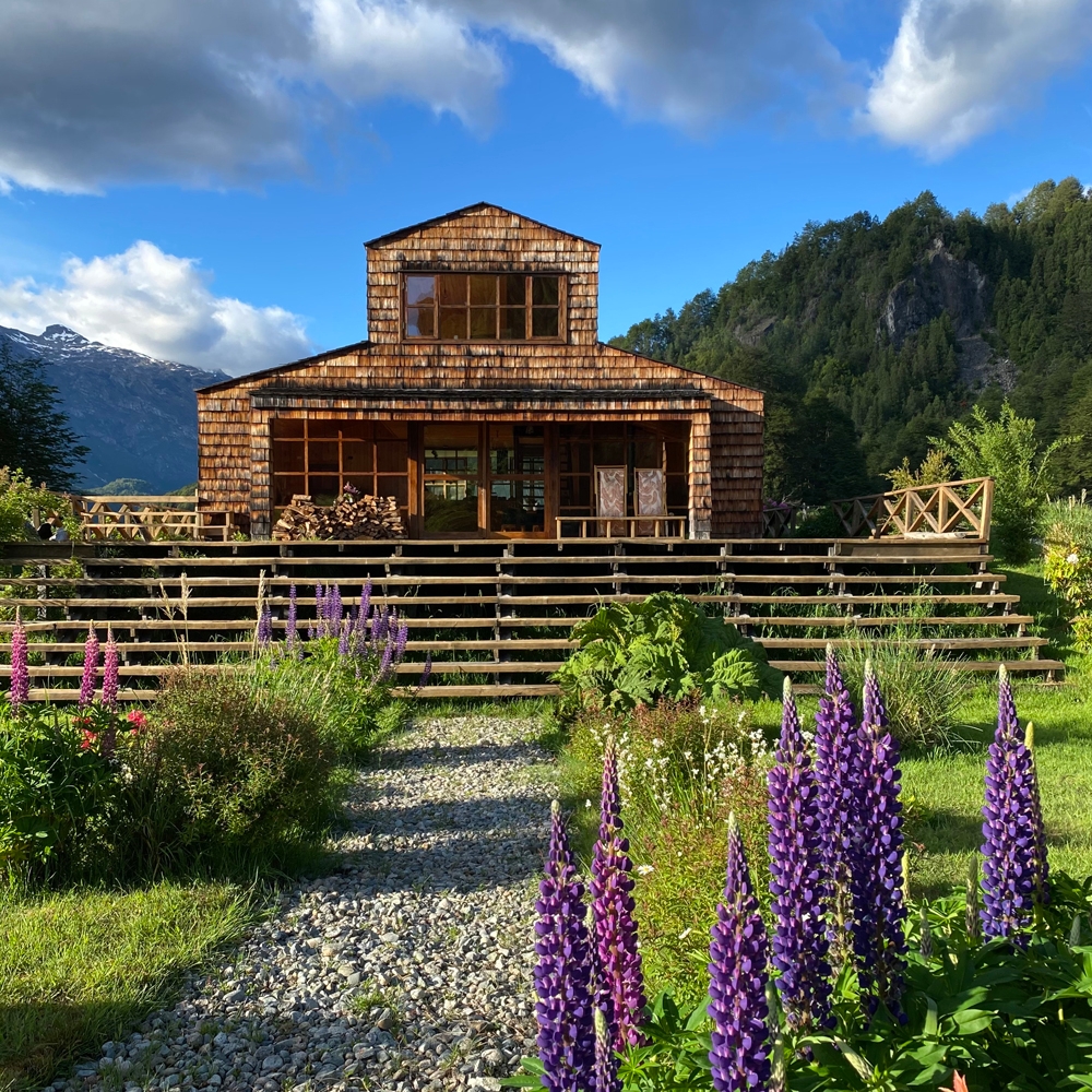 Wooden house with large windows, surrounded by a garden and mountains in the background.