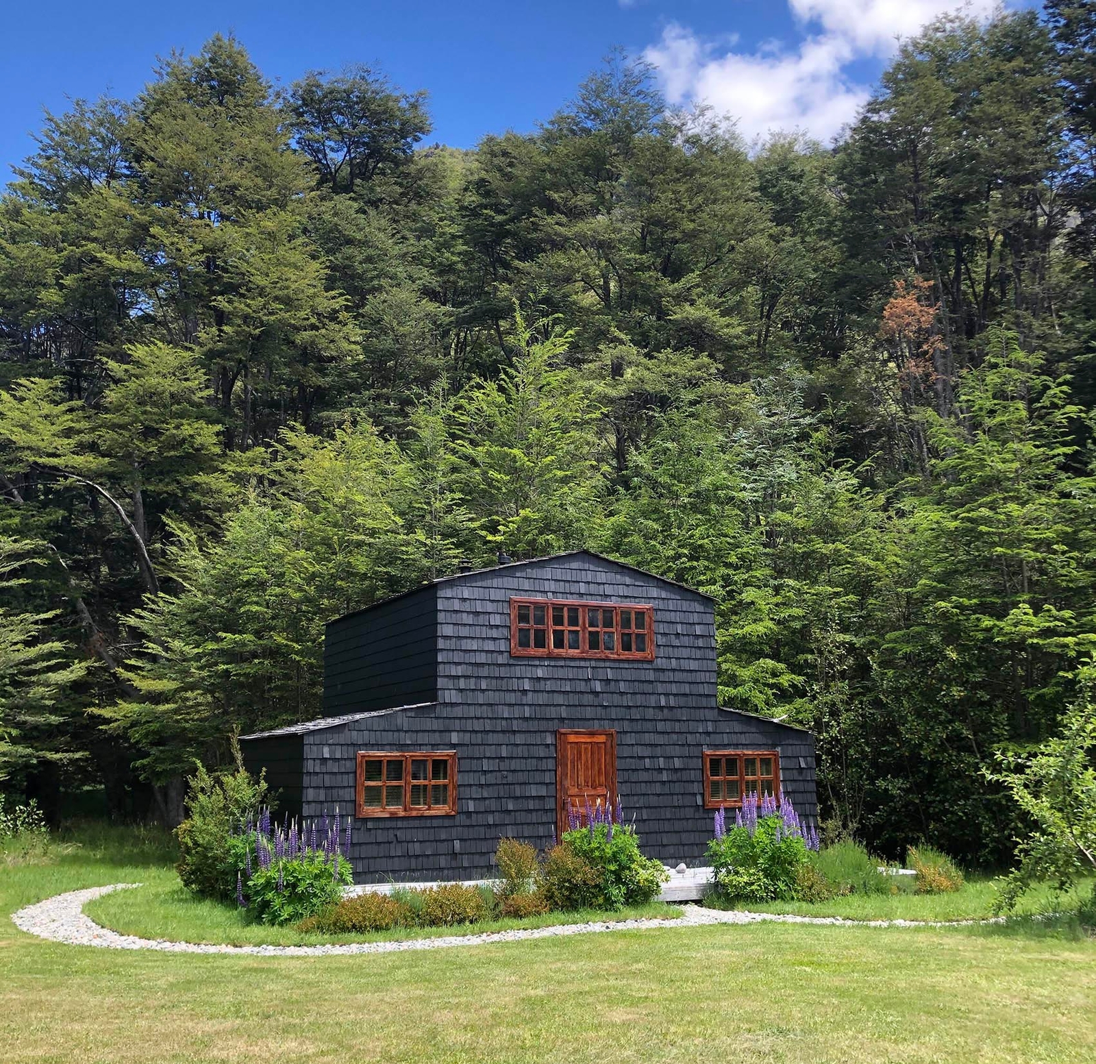 A two-story dark wooden cabin with a manicured lawn and forest backdrop under a blue sky.