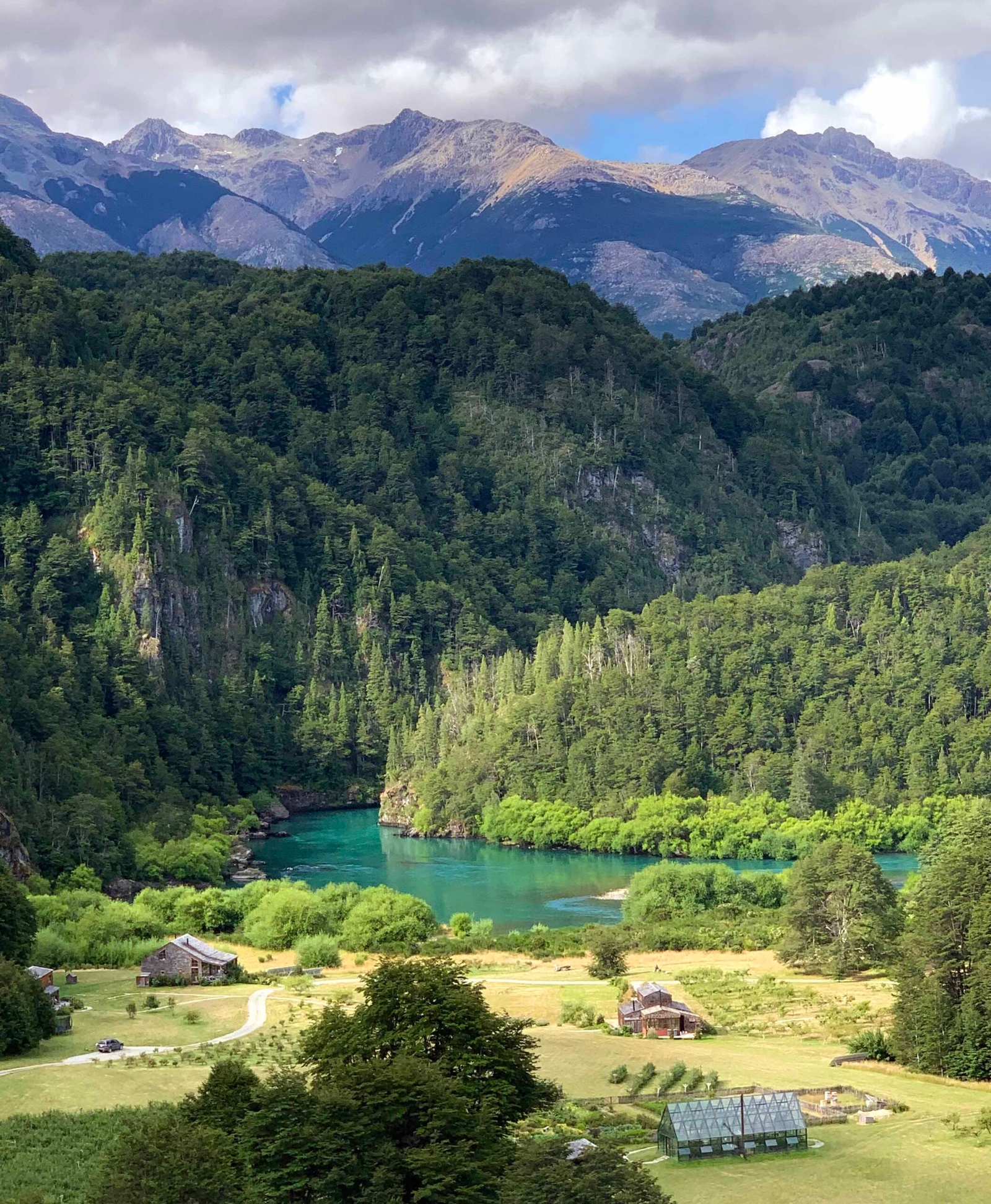 A lush valley with a clear blue river, surrounded by forests and mountains under a cloudy sky.