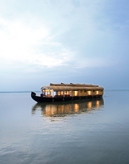 A houseboat with lit interiors floating on calm waters under a blue sky.