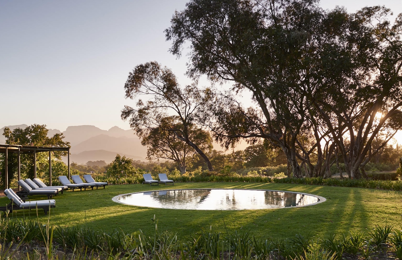 Round pool with lounge chairs in a serene garden at sunset, mountain backdrop.