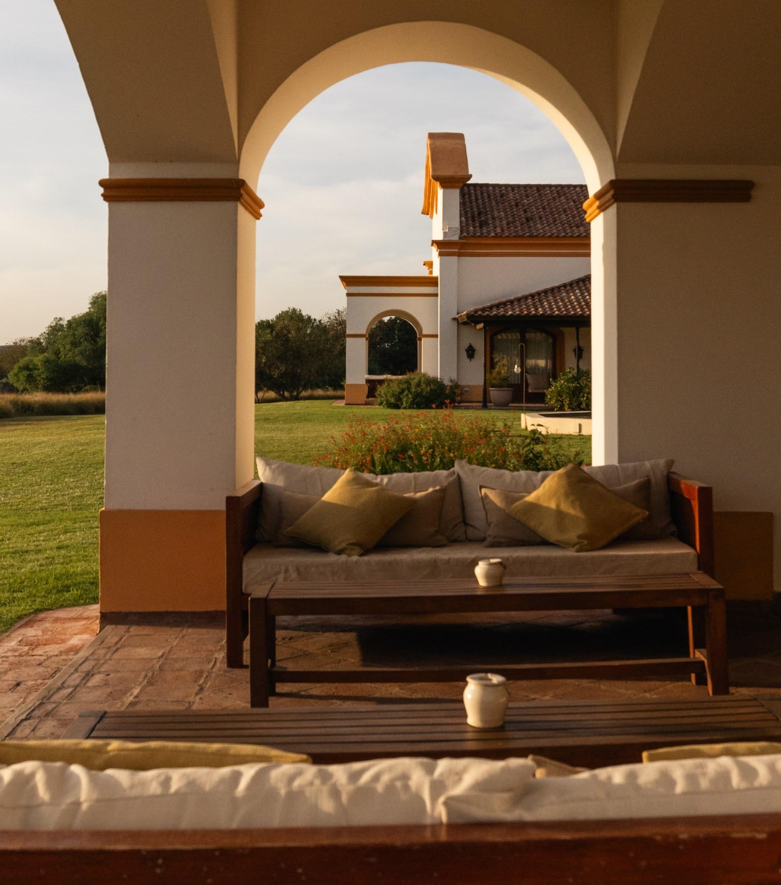 A cozy outdoor seating area with a wooden sofa and cushions, framed by arches overlooking a garden.